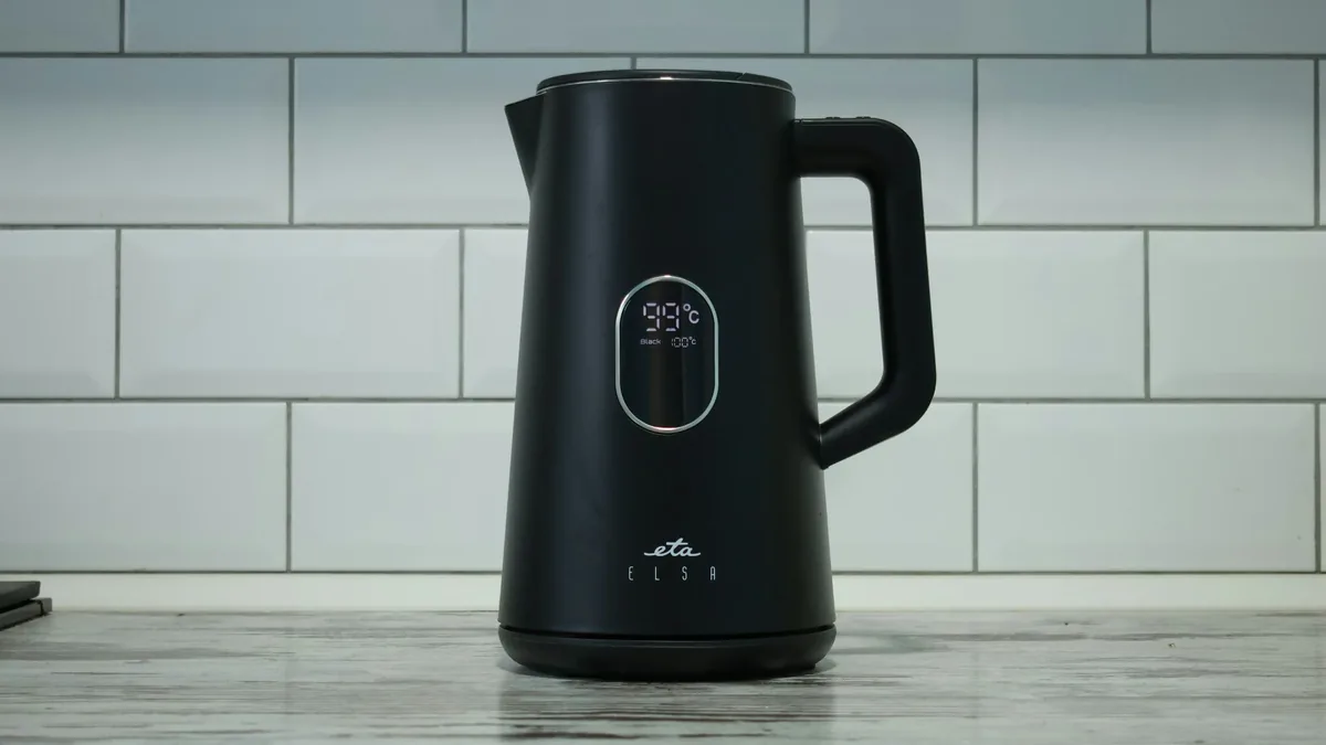 A black electric kettle with a digital display and a handle, sitting on a grey marble countertop in front of a white subway-tiled wall.