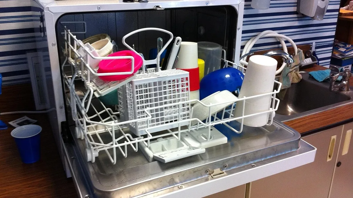 A dishwasher with its door open, revealing a rack containing various kitchen items, including dishes, cups, and utensils, situated on a coun