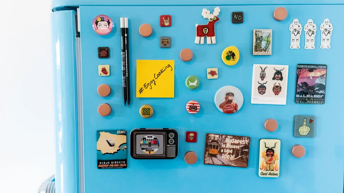 A light blue refrigerator with various magnets attached to its surface, including a yellow post-it note, a pen, and several small images.